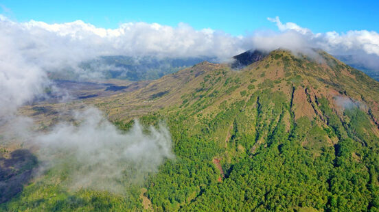 Aerial view of a lush green mountain ridge with white clouds clinging to its slopes under a clear blue sky.