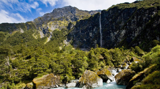 A long, slender waterfall falling from a high mountain cliff covered in green trees into a blue stream.