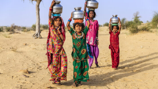 Group of young girls balancing metal water pots on their heads in a desert environment.