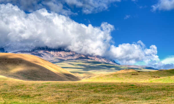 Sunlit rolling hills lead toward a massive mountain range capped with heavy white clouds under a bright blue sky.
