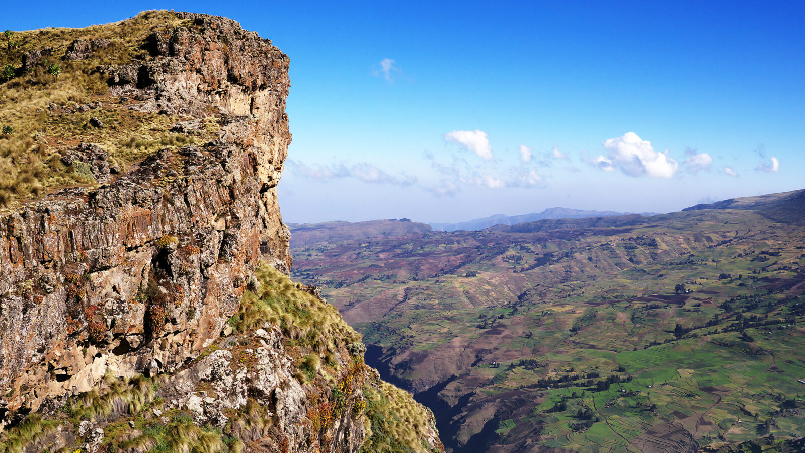 A rugged brown cliff face overlooks a massive valley with rolling green hills and distant mountain ranges.