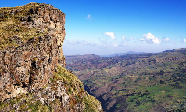 A rugged brown cliff face overlooks a massive valley with rolling green hills and distant mountain ranges.
