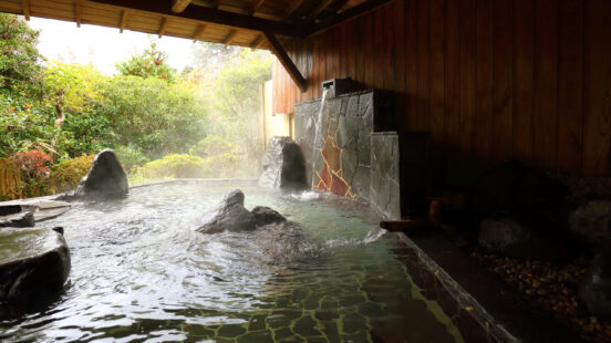 Steaming water in an outdoor stone hot spring pool under a wooden roof.