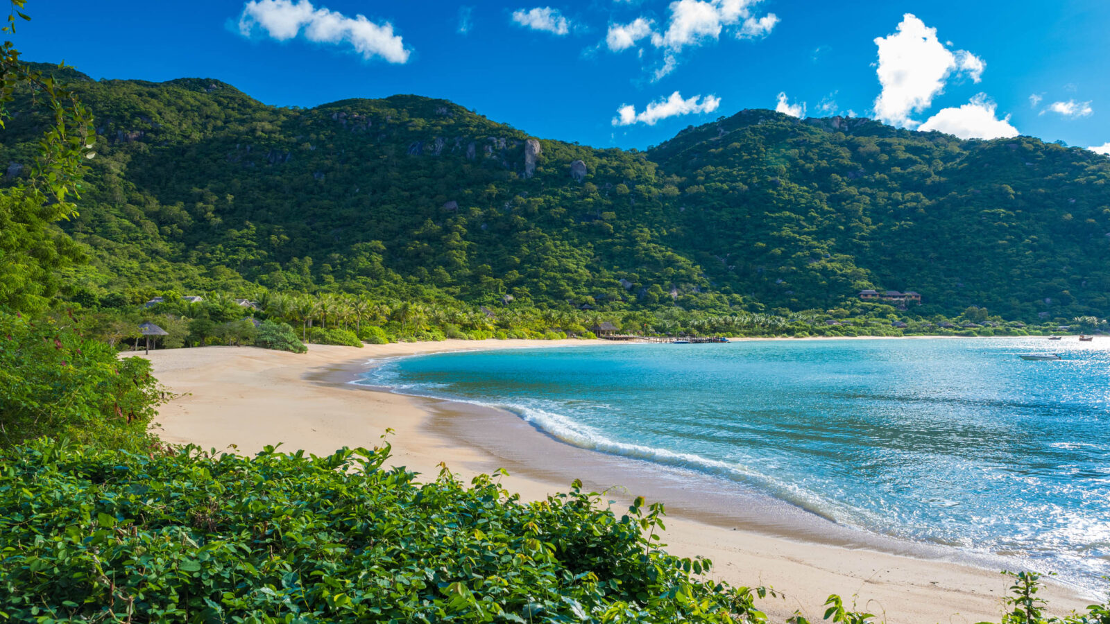 A curved sandy beach meets blue ocean water, bordered by lush green hills and a bright blue sky.