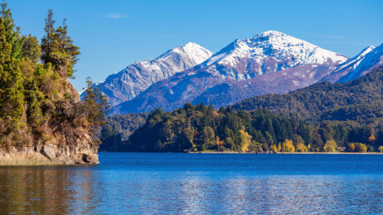 A blue lake in the foreground with evergreen trees and majestic snow-capped mountains under a clear blue sky.