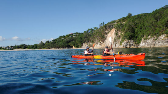wo people paddling a tandem orange kayak on calm blue water near a forested coastline.