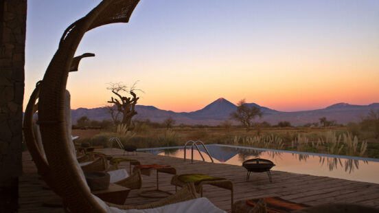 Poolside deck with wicker chairs overlooking a desert plain and a pointed volcano under a pink and orange sky.