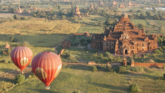 High-angle view of the Dhammayangyi Temple in Bagan with two red hot air balloons flying over green fields.