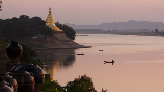 A golden pagoda sits on the bank of a wide river at dusk. Small boats row across the calm, pink-tinted water.