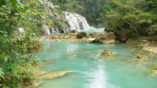 A wide turquoise river flows through a dense forest with small waterfalls and rocky banks.