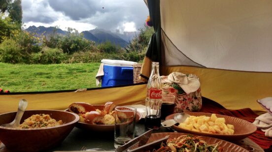 A breakfast or lunch spread inside a camping tent featuring bowls of food, a Coca-Cola bottle, and mountain views.