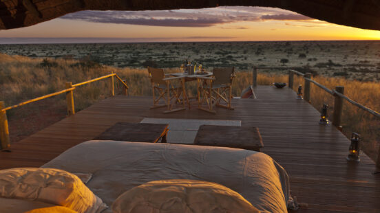 Luxury outdoor sleeping area and dining set on a wooden platform overlooking a grassy plain at sunset.