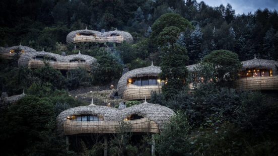 Several round thatched-roof buildings integrated into a dense, green forest on a hillside at dusk.
