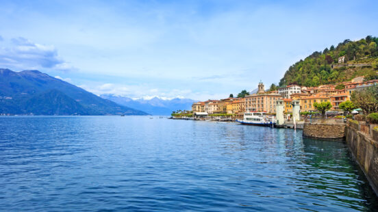 Wide shot of a lakeside town with colorful houses along the water and mountains in the distance.