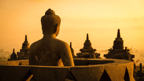 Stone Buddha statue seen from behind, looking out over stupas at Borobudur Temple during a golden sunset.