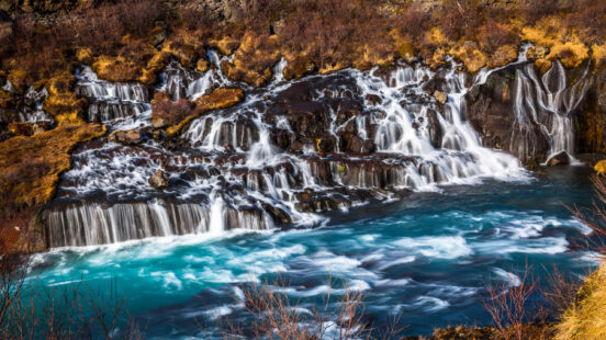 Numerous small waterfalls flowing over dark rocks into a vibrant blue river with orange and brown foliage.