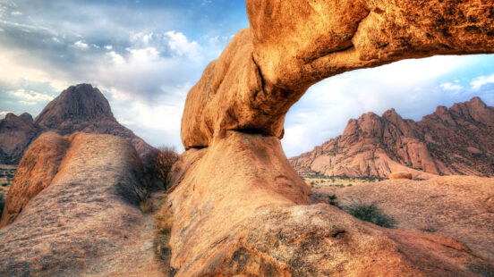 View of a massive orange rock formation with a natural bridge opening onto a mountain range.