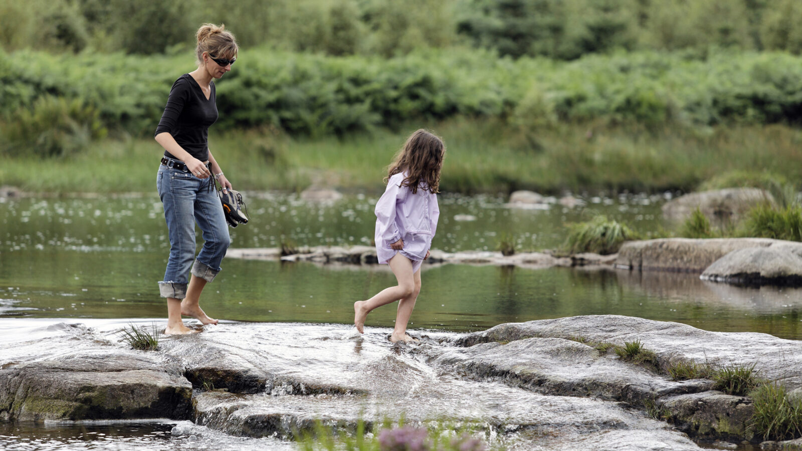 A woman and a young girl wade across a rocky stream in a green woodland setting.
