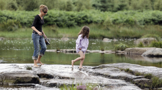 A woman and a young girl wade across a rocky stream in a green woodland setting.
