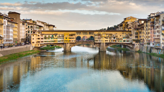 The Ponte Vecchio stone bridge with shops reflecting in the calm river water under a cloudy sky.