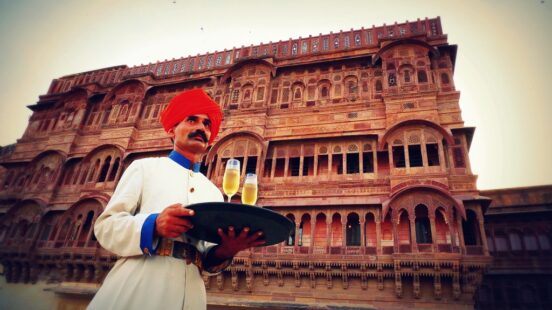 A man in a red turban holding a tray with two glasses in front of an ornate palace