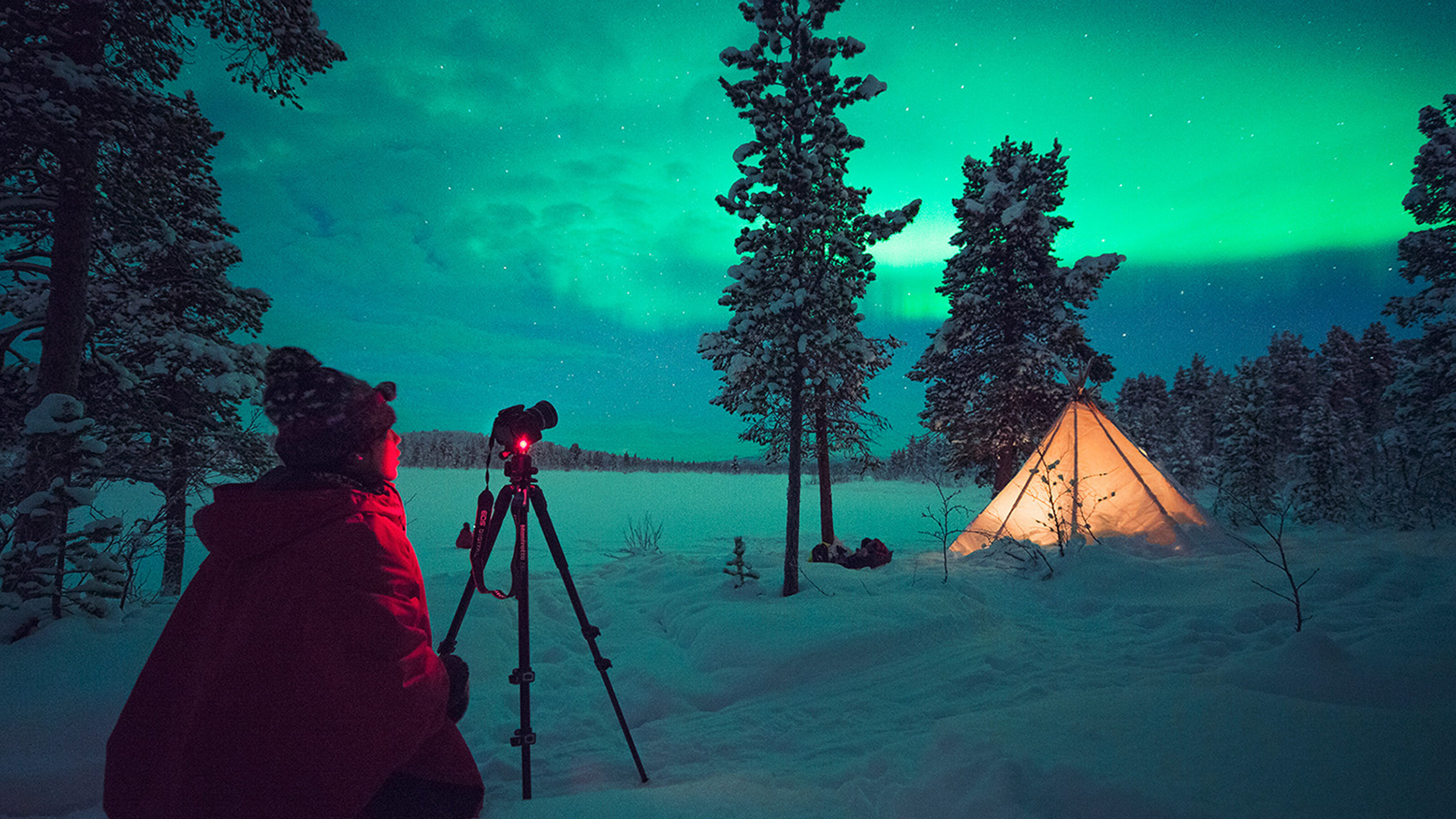 Person in a red jacket using a tripod-mounted camera to photograph green northern lights near a lit tent in the snow.