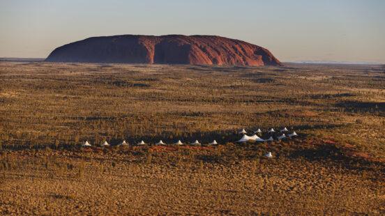 Wide shot of Uluru under a clear sky with several white pointed tents scattered across the desert scrub in the foreground.