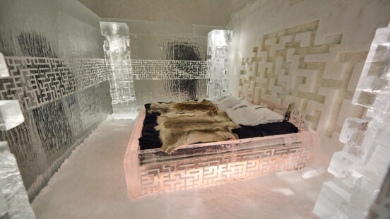 A bed with animal skins and white pillows inside a room with walls and furniture carved from blocks of clear ice.
