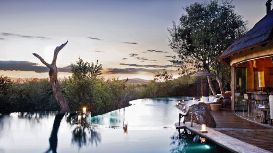 A tranquil infinity pool at dusk with candles on a wooden deck overlooking a dense thicket of green trees.
