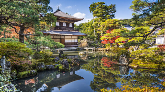 Traditional two-story wooden Japanese pavilion surrounded by manicured green moss, trees, and stone pathways.