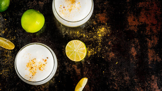 A high-angle view of two foamy white cocktails, limes, and lime slices on a dark, rustic surface.