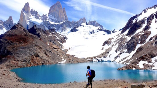 A hiker with a backpack and poles stands on the shore of a bright turquoise glacial lake looking toward the dramatic snow-capped peaks of Fitz Roy.