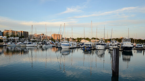 A rows of luxury yachts and boats docked at a marina during sunset with city buildings behind.