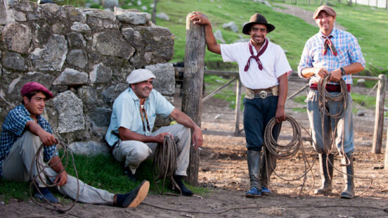 Four smiling men in traditional gaucho attire sit and stand outdoors, holding coils of thick rope.