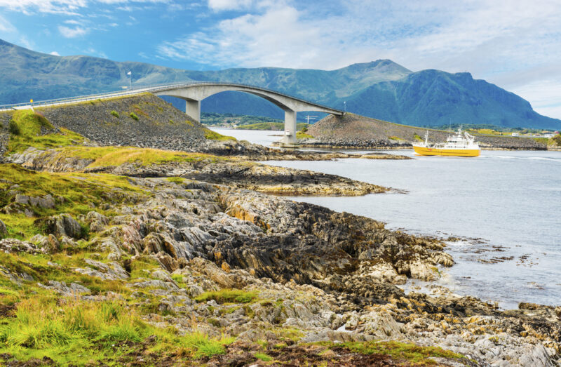 A curved bridge spans across the sea between rocky shores with a yellow ship passing underneath.