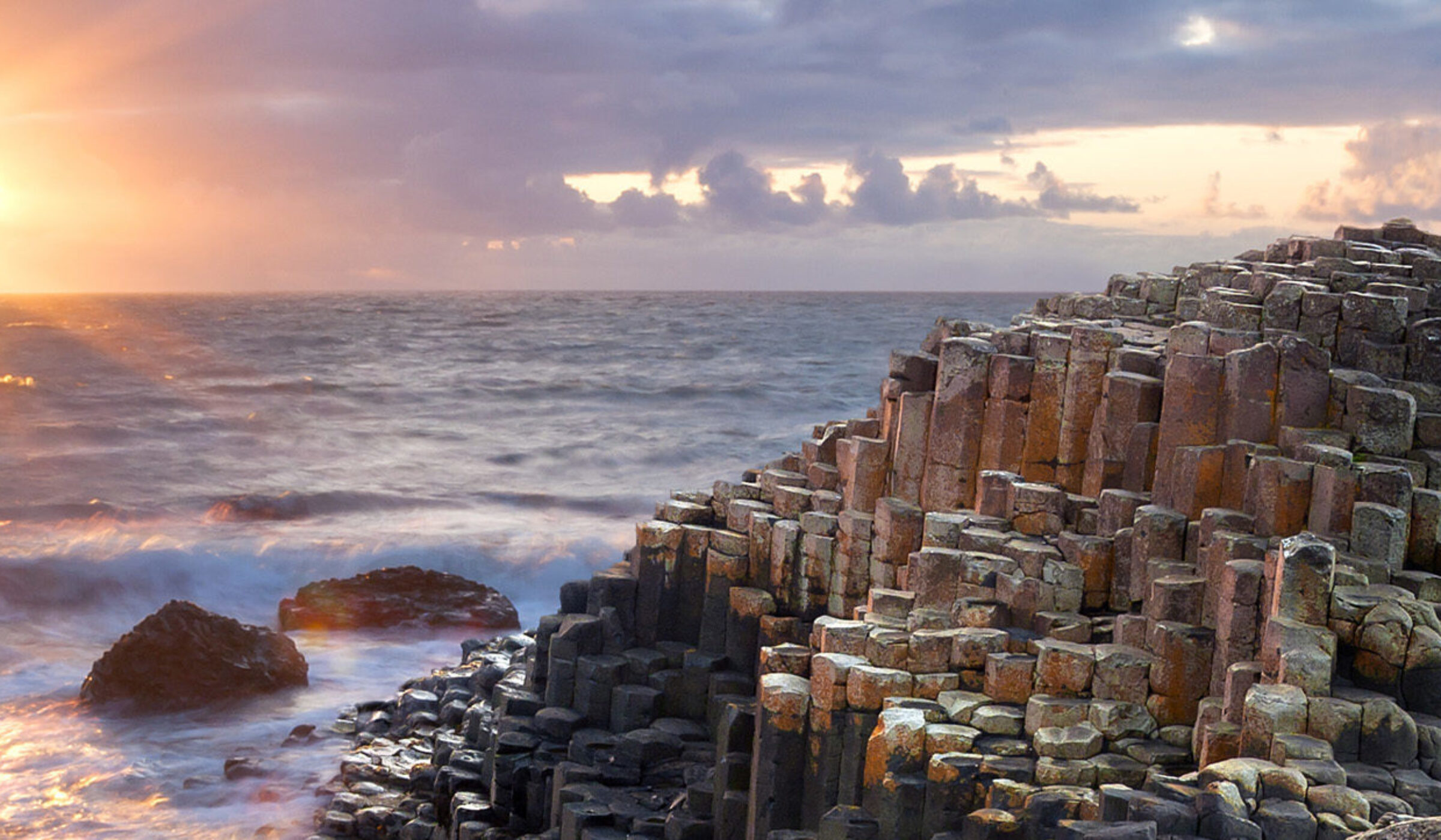 Panoramic photo of Giant's Causeway basalt columns at sunset, with golden light reflecting on the ocean water. Book your luxury Ireland tour.