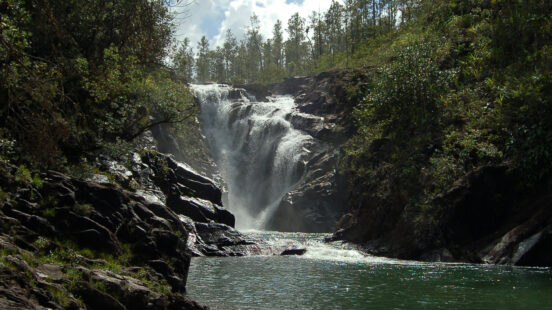 A large waterfall flowing into a river basin surrounded by rocky cliffs and green trees.
