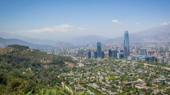 A wide cityscape of Santiago, Chile, with skyscrapers and surrounding houses beneath distant mountains.