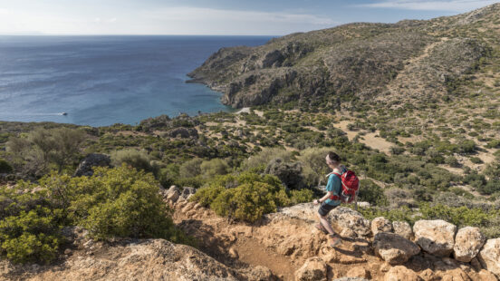 A person hiking on a rocky trail with a scenic view of the blue ocean and dry, scrub-covered hills.