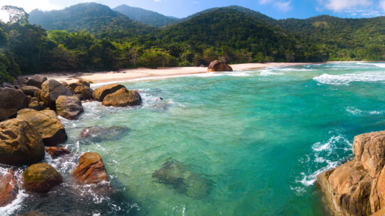 Large brown boulders in the foreground of a tropical beach with turquoise water and forested mountains.