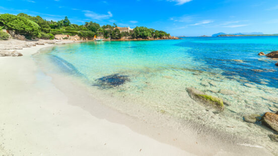 Transparent blue water on a sandy shore with green trees and rocks in the background.