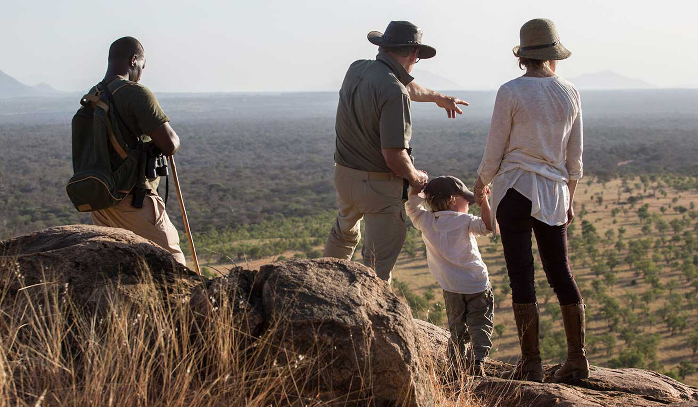 adventure trips - Four people wearing neutral clothes on safari in Kenya - an adult man and woman hold the hands of a young child in between them and point out at the landscape. Another adult man rests against a large rock.