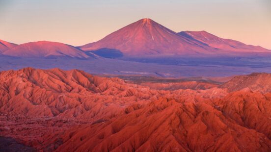 A wide shot of the Atacama Desert showing red jagged hills in the foreground and a purple volcano at sunset.