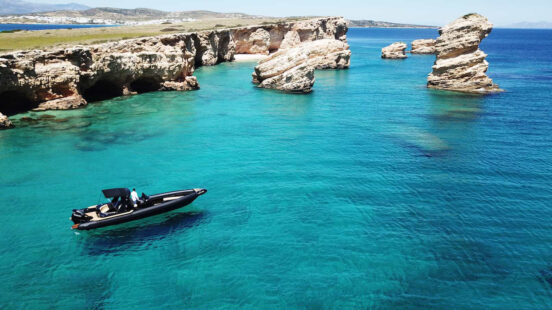 A black RIB boat navigating crystal clear turquoise water by rocky island cliffs.