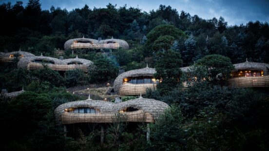 Several round thatched-roof lodges nestled amongst dense green trees on a steep hill during twilight.
