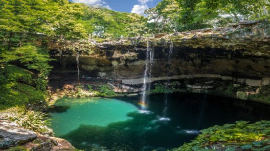 A waterfall drops into a turquoise pool within a rocky cenote cave surrounded by vibrant green trees and plants.