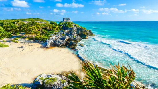 Ancient stone ruins on a cliff overlooking a tropical white sand beach and turquoise ocean waves under a blue sky.