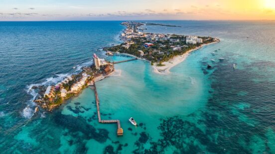 Aerial view of a thin tropical island with turquoise water, resorts, and a long wooden pier extending into the sea.