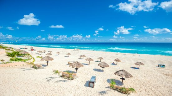 Wide shot of a sunny white sand beach with several thatched umbrellas and bright turquoise Caribbean-style water.
