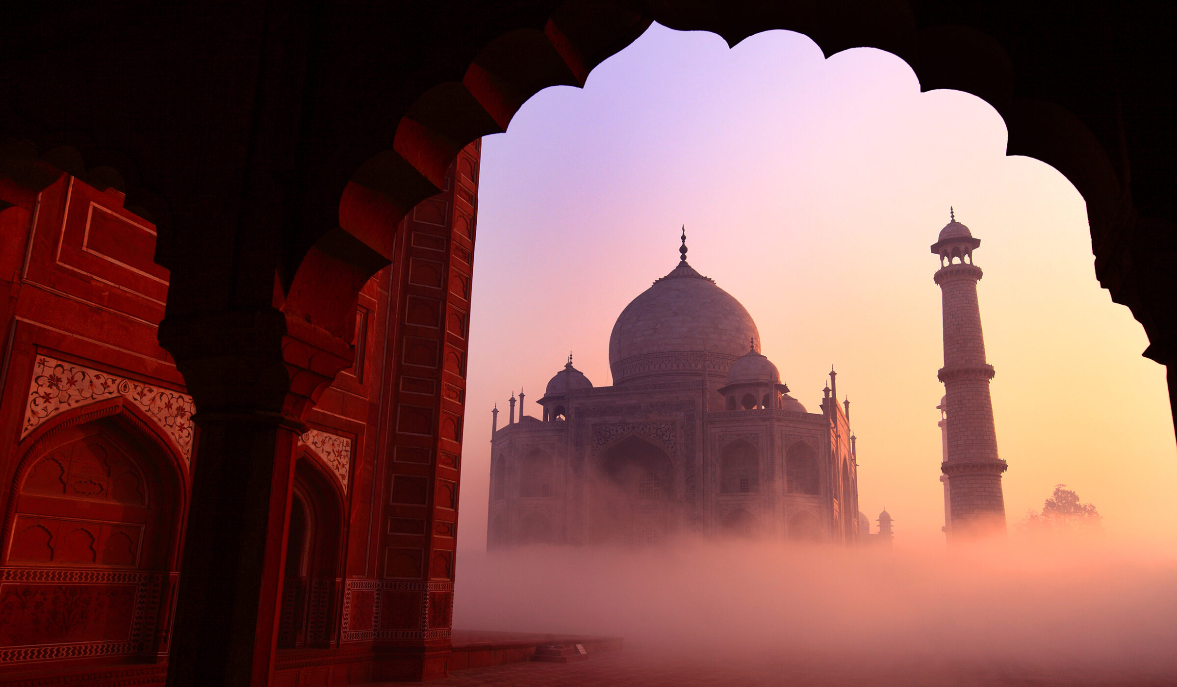 The Taj Mahal shrouded in pink morning fog viewed through an archway during luxury history tours.